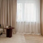 Jasny salon w mieszkaniu na sprzedaż w Opolu z beżową kanapą i oknem z firanami